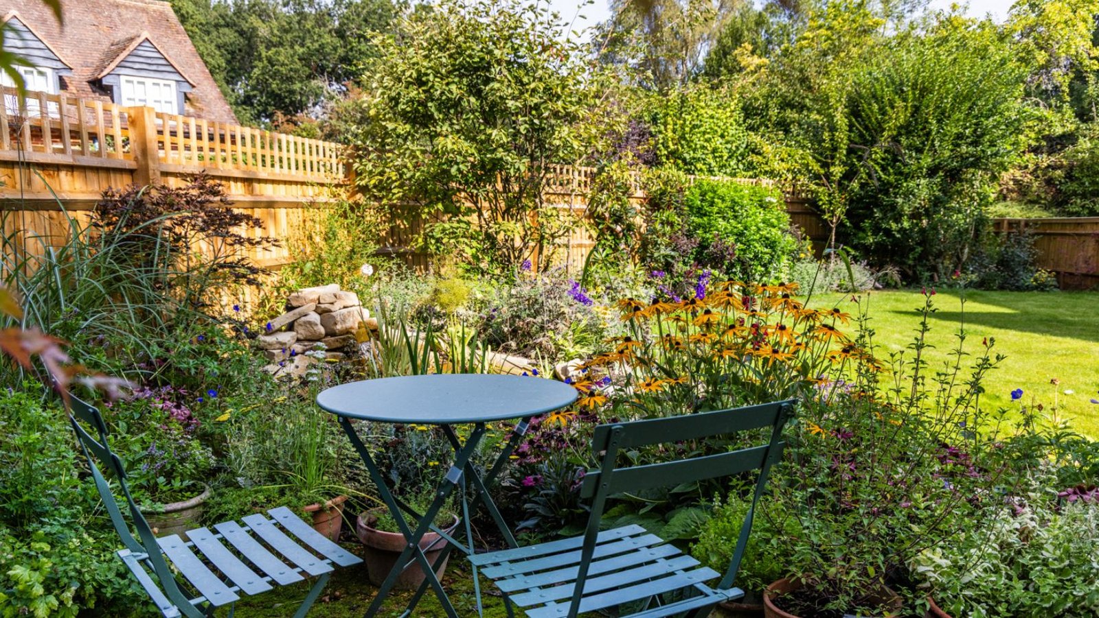 Image of bistro garden set, surrounded by garden beds filled with flowering plants and a water feature